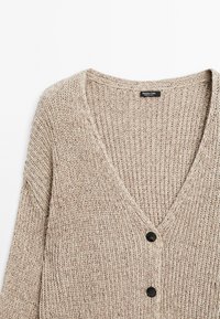 Beige stickad kavaj med V-ringning, prydd med ett strukturerat mönster och tre svarta knappar fram. Lätt och avslappnad passform.