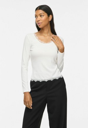 Femme aux longs cheveux foncés portant un haut blanc à manches longues avec bordure en dentelle et un pantalon noir, posant avec une main près du cou.