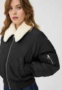 Czarna kurtka bomberka z kremowym sztucznym futrem przy kołnierzu, bocznymi kieszeniami oraz kieszenią na ramieniu zapinaną na zamek. Posiada zamek błyskawiczny z przodu i ściągacze przy mankietach.