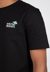 Camiseta de algodón negra con un pequeño logotipo blanco que dice "WRONG FRIENDS" y un diseño en acento verde. Mangas cortas y cuello redondo.