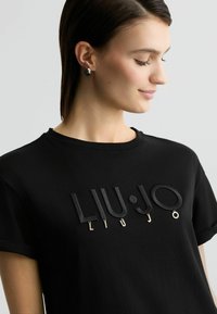 Must üleüldine T-särk, millel on reljeefne, tekstuuriga "LIU•JO" logo erinevates kõrgustes ja kullatooni aktsendid. Klassikaline ümar kaelus ja lühikesed varrukad.