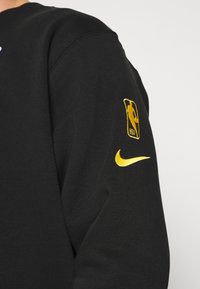 Svart sweatshirt med en NBA-logopatch i gult på vänster ärm och en gul Nike-swoosh på höger ärm. Mjuk textur.