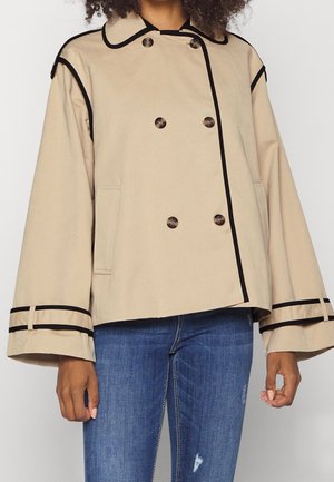 Kvinde iført beige dobbeltradet jakke med sort kant og vide ærmer, kombineret med blå slidte jeans.