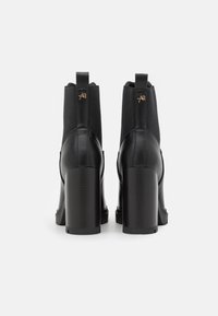 Mustat nilkkurit, joissa korkeat korot, sileää nahkaa olevat paneelit ja joustavat sivut. Kenkässä on nelikulmainen korko ja pieni kohokuvioitu logo takana.