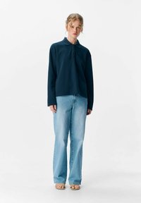 Polo shirt a maniche lunghe blu navy con colletto e tre bottoni, abbinato a jeans chiari a campana e sandali chiari.