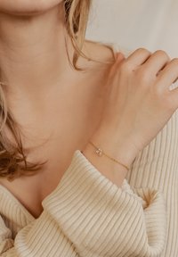 Gold bracelet with a delicate chain and a floral charm featuring a small crystal accent. The hand rests against a textured beige sweater.