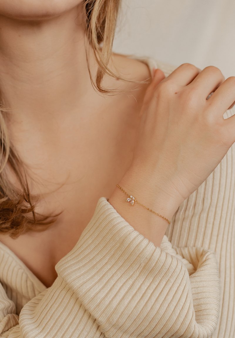 Gold bracelet with a delicate chain and a floral charm featuring a small crystal accent. The hand rests against a textured beige sweater.
