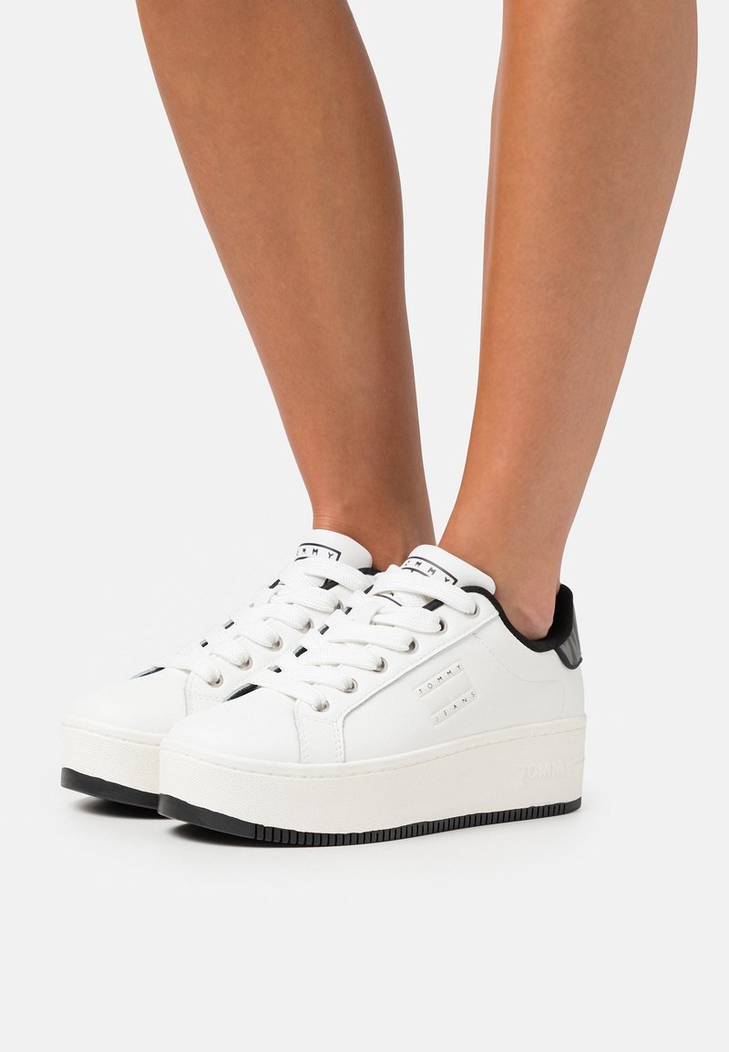 Baskets blanches à plateforme avec accents noirs au talon et lacets blancs portées sur des jambes nues sur un fond clair.
