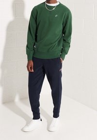 Zelená mikina s kulatým výstřihem, texturovaná tkanina, malý detail loga. V kombinaci s námořnickými joggery a bílými teniskami, na světlém pozadí.