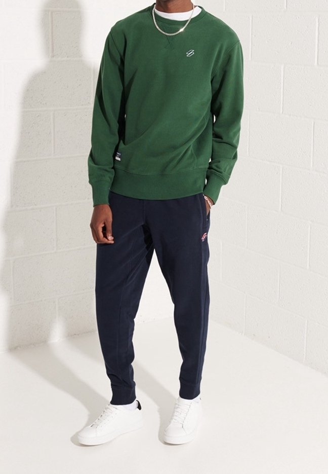 Zelená mikina s kulatým výstřihem, texturovaná tkanina, malý detail loga. V kombinaci s námořnickými joggery a bílými teniskami, na světlém pozadí.