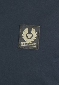 Tengerészkék szövet, fekete textúrájú folttal, amelyen egy arany embléma és a "BELSTAFF" név látható alul.