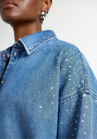 Camicia in denim blu caratterizzata da un colletto classico, abbellita con pezzi decorativi lucidi sulle spalle, che aggiungono texture e interesse visivo.