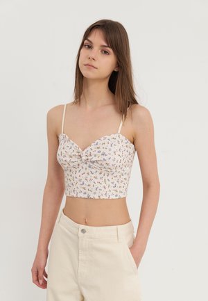 Top corto floral con escote en forma de corazón, fondo blanco y patrones de flores multicolores. Presenta tirantes finos y un detalle de torsión en la parte frontal.