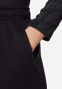 Prenda deportiva negra con un bolsillo y mangas texturizadas. El material parece suave con un acabado liso. Los acentos incluyen un ajuste entallado y una cinturilla elástica.