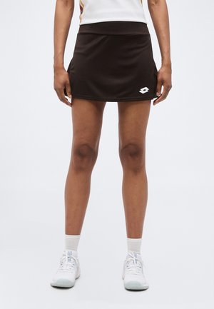 Žena nosí čiernu športovú sukňu s logom na ľavej strane, biele ponožky a biele športové topánky, stojí čelom dopredu.