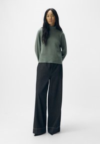 Groene coltrui van zacht materiaal, gecombineerd met zwarte broek met wijde pijpen en een streepjesmotief met contrasterende stiksels.