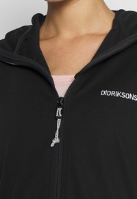 Černá zipová bunda s měkkou texturou, vybavená kapucí, elastickou šňůrkou a vyšitým logem značky na hrudi.