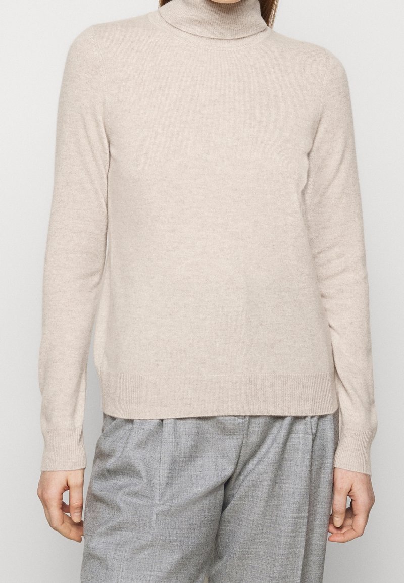 Pull à col montant beige clair, fabriqué en tricot doux. Design ajusté avec des manches longues et un ourlet côtelé, associé à un pantalon gris.