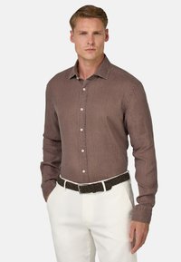 Camicia a maniche lunghe marrone con un motivo testurizzato, colletto alla francese e bottoni bianchi, indossata con una cintura intrecciata marrone e pantaloni bianchi.
