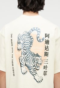 Πίσω μέρος λευκού πουκαμίσου με γραφικό τίγρης σε ασπρόμαυρο, πορτοκαλί κυκλικό φόντο και μαύρο κείμενο στα αγγλικά και κινέζικα.