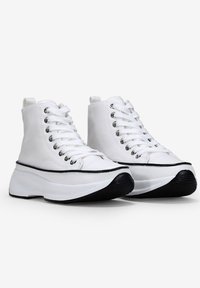 Sneakers alti bianchi con suola nera, in materiale tela, punta rotonda e un sistema di allacciatura ben evidente. Presentano una striscia nera a contrasto lungo la suola.