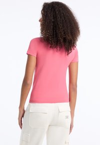 Trumpas rankovėmis, prigludęs marškinėliai ryškiai rožinės spalvos medvilnės audinio. Atvirkštinis vaizdas rodo lygų tekstūrą ir paprastą dizainą, derinamas su smėlio spalvos cargo kelnėmis.