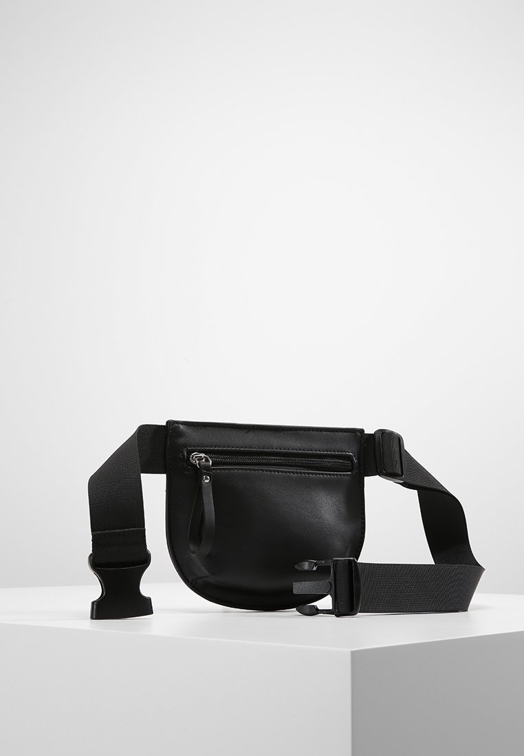 Borsa a vita nera in pelle con tasca frontale con zip e cinturino regolabile in tessuto nero con chiusura in plastica, esposta su una superficie bianca.