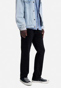 Lichtblauwe denimjas over een donkerblauw shirt, gecombineerd met zwarte broek en zwarte sneakers. Kenmerken zijn onder andere knopen en contrasterende texturen.