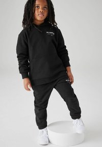 Set di felpa nera con cappuccio e pantaloni tuta, caratterizzato da una tasca frontale e polsini elasticizzati, completato da dettagli di sneakers bianche. Logo stampato su entrambi i pezzi.
