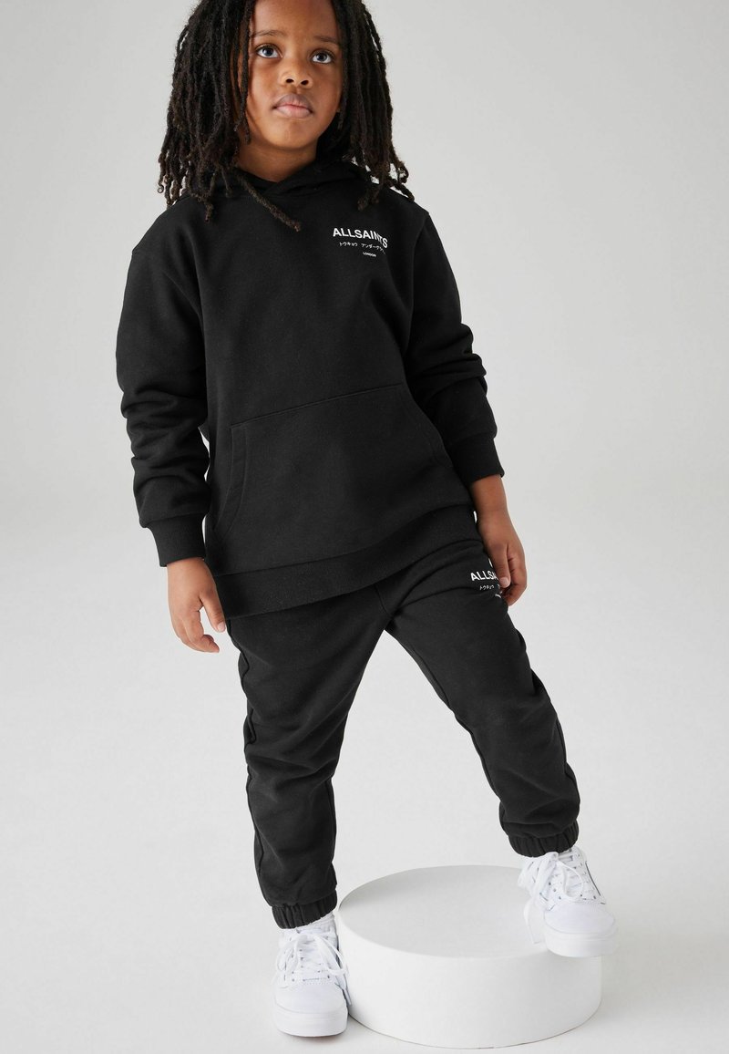 Set di felpa nera con cappuccio e pantaloni tuta, caratterizzato da una tasca frontale e polsini elasticizzati, completato da dettagli di sneakers bianche. Logo stampato su entrambi i pezzi.