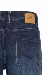 Jean en denim bleu marine avec un patch en cuir portant "camel active". Comprend deux poches arrière avec des détails de couture.