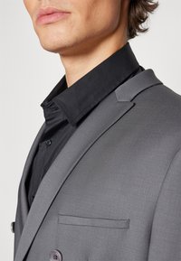 Grå skreddersydd blazer med notklap, med en enkelt lomme og dobbeltknapping. Under er en svart skjorte synlig.