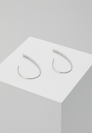Par tynde, sølvfarvede, åbne hoop-øreringe med et minimalistisk buet design placeret på en hvid piedestal mod en grå baggrund.