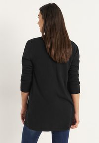Frau mit langen braunen Haaren dreht sich um, trägt einen schwarzen langen Pullover und blaue Jeans vor einem einfarbigen hellen Hintergrund.