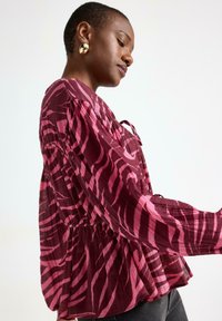 Kohokulta-bluusi tummanpunaisena ja vaaleanpunaisena, jossa on seeprahkuvio, pitkähihaiset hihat, kootut vyötärö ja nauhakiinnitys kaula-aukoissa.