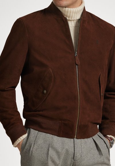 Rjava semiš bomber jakna z rebrastimi manšetami, stranskimi žepi in zapiranjem na zadrgo. Oblečena preko kremnega puli ovratnika. Vidni minimalistični detajl blagovne znamke.