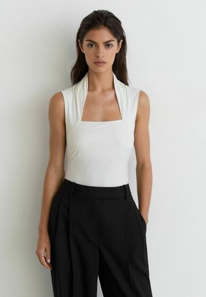 Femme aux longs cheveux foncés portant un haut blanc sans manches avec un décolleté carré et un pantalon noir taille haute, debout contre un mur blanc.