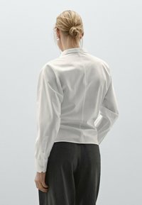 Blusa bianca a maniche lunghe con design aderente, dettagli sulla cucitura posteriore e polsini con bottoni, abbinata a pantaloni grigi strutturati.