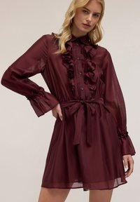 Abito camicia color borgogna realizzato in tessuto leggero e trasparente. Presenta un davanti arricciato, chiusura con bottoni e vita legata. Maniche lunghe e arricciate.