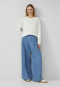 Vrouw die tegen een muur staat, gekleed in een witte trui, brede blauwe streepje jeans en witte sneakers met rubberen zolen.