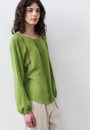 Femme aux cheveux bouclés et foncés portant un chemisier vert à manches longues avec des détails en dentelle et un pantalon clair à motifs, debout devant un fond blanc.