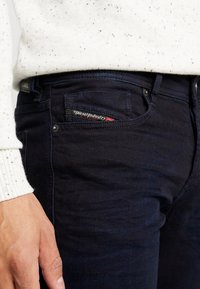 Los jeans de denim de color azul oscuro presentan un bolsillo frontal con una etiqueta de Diesel Industry. La tela tiene una textura suave y un aspecto ligeramente desgastado.