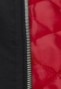 Schwarzer Stoff mit einem silbernen Reißverschluss auf einer Seite und einem roten gepolsterten Innenfutter auf der anderen. Das Muster zeigt diamantförmige Formen.