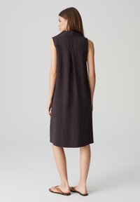 Mouwloze donkergrijze linnen jurk met een kraag, plooi detail aan de achterkant en een lengte tot de knie. Draagt een paar bruine flip-flop sandalen.