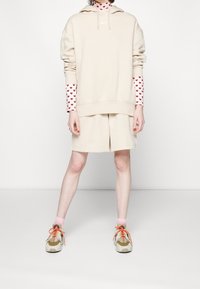 Sweat à capuche beige avec un logo blanc, assorti à un short assorti. En dessous, un tee-shirt à manches longues à motifs avec des cœurs rouges. Baskets beige et orange.