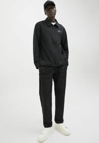 Schwarze Zip-Up-Jacke mit Kragen und Logo auf der Brust, kombiniert mit schwarzen Cargo-Hosen mit Taschen und weißen Sneakers.