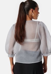 Blusa grigia chiara trasparente con maniche a sbuffo e finitura testurizzata. La vista posteriore mostra un design sottile e tessuti a strati.