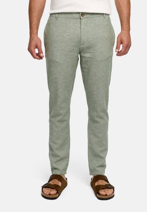 Mężczyzna ubrany w jasnozielone dopasowane spodnie, białą koszulę i brązowe sandały na klamrę, stojący na białym tle.