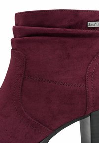 Bottine en suède bordeaux avec un col superposé, un talon noir élégant, des détails de couture et une petite étiquette métallique sur le côté.