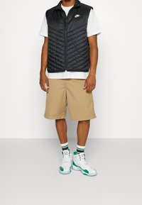 Svart vadderad väst med hög krage, vit t-shirt under, beigefärgade shorts och vit-gröna sneakers med mönstrade strumpor.